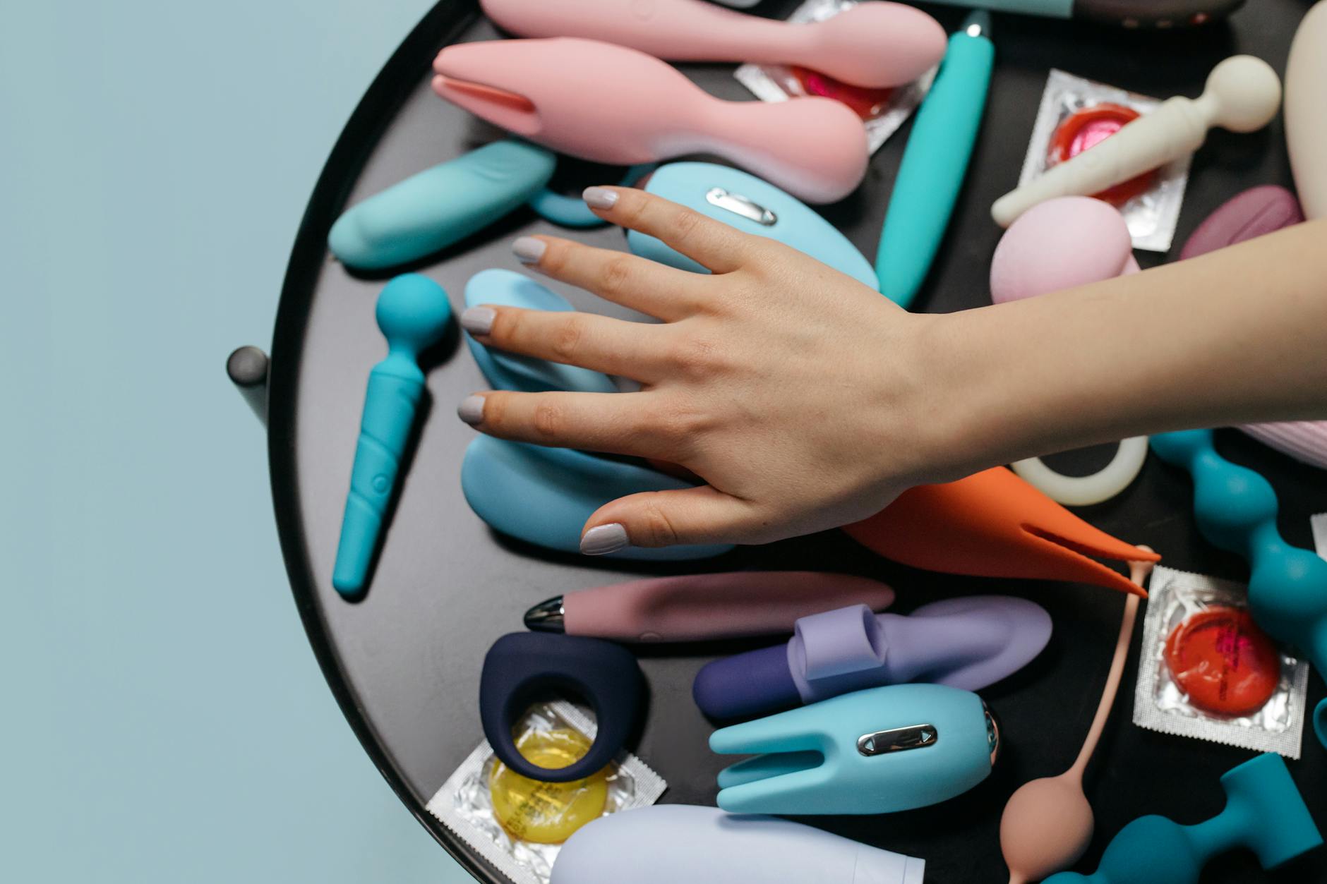 A hand reaching over a variety of colorful silicone vibrators arranged on a table.