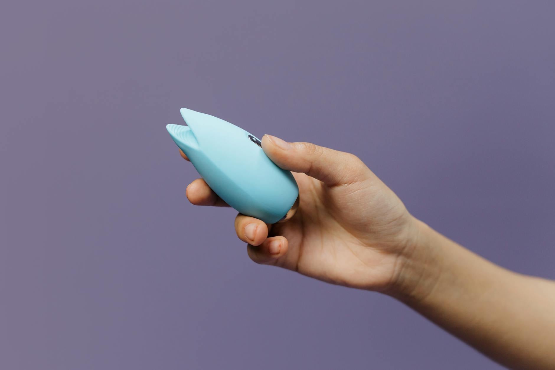 A blue silicone clitoral vibrator held in hand against a purple background, promoting self-love and sexual wellness