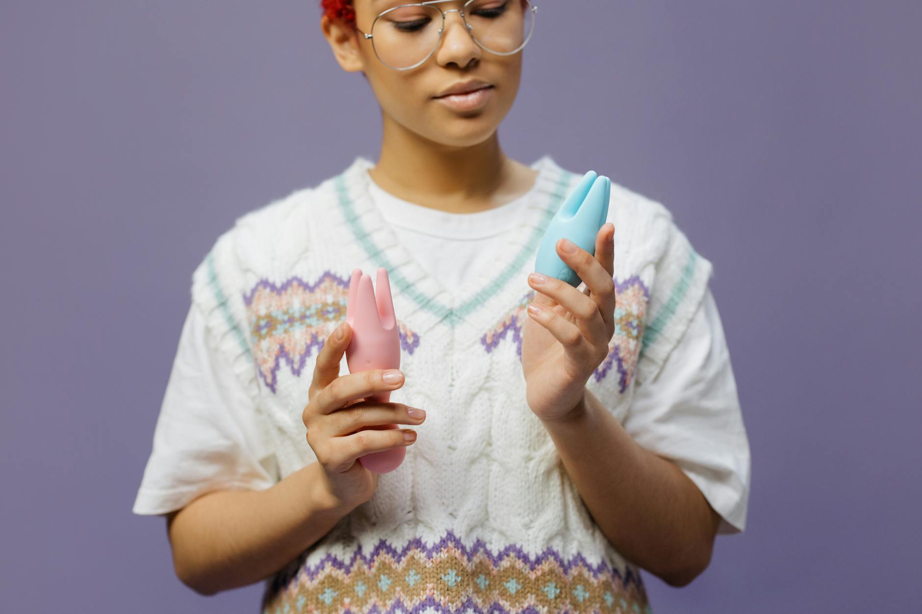 Two hands holding pink and blue silicone vibrators against a pastel background, representing couple exploration and shared intimacy.